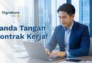 Seorang profesional atau karyawan baru mengenakan jas biru sedang tersenyum meninjau dan melakukan tanda tangan kontrak kerja digital melalui laptop di ruang kantor.