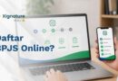 Seseorang sedang mengakses panduan alur pendaftaran BPJS online melalui layar laptop dan aplikasi smartphone di atas meja kerja kantor.